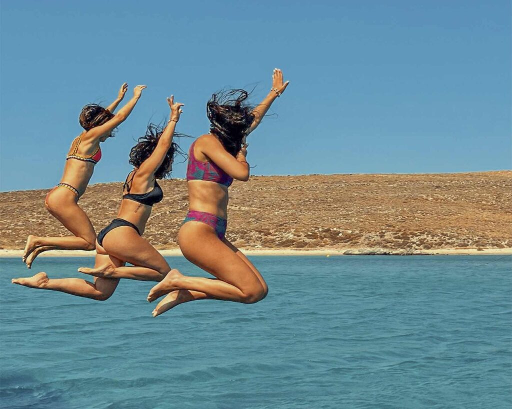 Three friends jumping into the crystal clear Aegean from a Don Blue Yachting private boat — the kind of spontaneous moment that makes the Paros vs Mykonos debate irrelevant