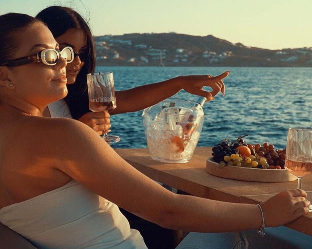 Two friends enjoying a sunset aperitivo on board a Don Blue Yachting private boat with the Aegean as their backdrop — the kind of moment that makes any time the best time to visit Mykonos
