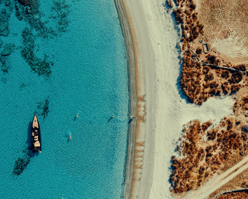 Aerial view of a secluded beach in the Cyclades with a private Don Blue Yachting boat anchored in crystal clear water — the real Paros vs Mykonos experience